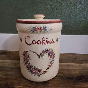 May include: A cylindrical ceramic cookie jar with a speckled cream finish, featuring a hand-painted floral heart motif and the word Cookies in red script. The jar includes a matching lid with a knob handle and red-painted trim along the edges.