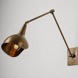 Può includere: Una lampada da parete con finitura in ottone anticato, dotata di una piastra di base quadrata, un braccio articolato regolabile con due punti di snodo e un ampio paralume a forma di campana che ospita una lampadina a filamento a vista.
