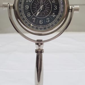 May include: A silver-toned metal desk clock featuring a circular face with a black background, white Arabic numerals, and intricate white patterns. The clock is mounted on a swiveling gimbal frame supported by a slender pedestal and a circular base.