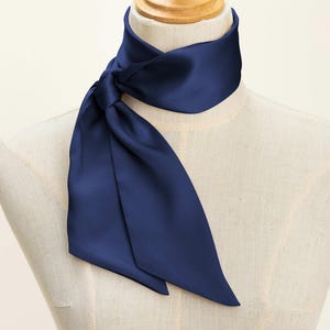 May include: A solid navy blue neck scarf tied in a simple knot around the neck of a mannequin. The scarf features a smooth, satin-like finish with a slight sheen and pointed ends that drape downward, creating a clean and polished appearance.