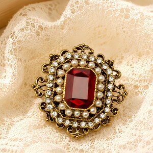 May include: A rectangular brooch featuring a large, deep red faceted center stone surrounded by a border of small white pearls. The frame is made of antiqued gold-tone metal with intricate scrollwork and a outer border of clear crystals.