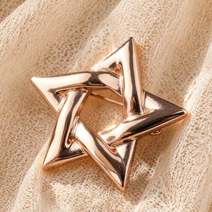May include: A polished rose gold-tone metal brooch shaped like a six-pointed star. The design features interlocking geometric bars with a smooth, reflective surface that catches the light, creating a clean and structured appearance for this accessory.