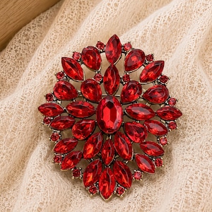 May include: A circular brooch featuring a large central oval red gemstone surrounded by multiple layers of marquise and pear-shaped red crystals set in an antique gold-toned metal frame with small round red accent stones throughout the design.