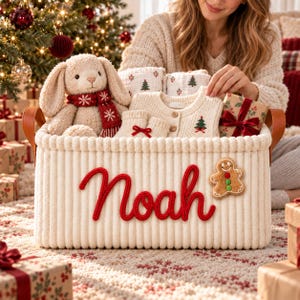 May include: A cream-coloured, ribbed storage basket with brown leather handles, featuring the name Noah in red script. Inside are a plush bunny with a red scarf, a cream knit cardigan with a small tree, and a gingerbread man patch on the front.