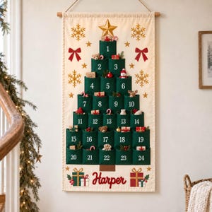May include: A cream-colored fabric wall hanging featuring a green felt Christmas tree shape with twenty-four numbered pockets. The design includes gold stars, snowflakes, red bows, and the name Harper in red script above two gift box illustrations.