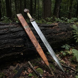 Könnte beinhalten: Ein zweischneidiges Gladius-Schwert mit einer polierten Stahlklinge, einem hellen Griff und einem dunklen Knauf. Das Schwert wird von einer braunen Lederscheide begleitet, die mit komplexen Prägemustern und Metallakzenten versehen ist.