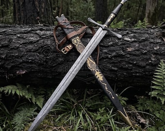 May include: A longsword with a patterned steel blade, a black grip, and a crossguard. The sword is accompanied by a black scabbard featuring gold-colored leaf and vine filigree, brown leather straps, and a metal chape at the pointed end.