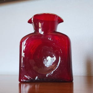May include: A translucent ruby red glass pitcher with a square-shaped body, a short neck, and a wide, flared spout. The glass features a smooth, reflective surface with a circular indentation centered on the front face of the vessel.