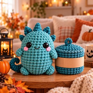 Puede incluir: Un juguete de peluche de dinosaurio tejido a ganchillo en color verde azulado con grandes ojos negros y mejillas rosadas, junto a un tarro tejido a ganchillo con tapa a juego envuelto en una banda de papel color canela.