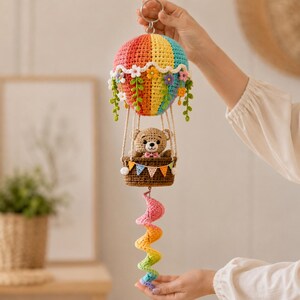 May include: A crocheted hot air balloon ornament featuring a multi-colored striped balloon canopy adorned with small fabric flowers and green vines. A brown crocheted teddy bear sits in the basket, which is decorated with a colorful triangular bunting.