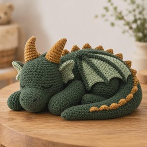 May include: A crocheted dragon toy in a sleeping position, featuring a dark green body, light green wings, and mustard yellow horns and back spikes. The creature has closed eyes and a textured yarn surface, crafted with detailed stitching.