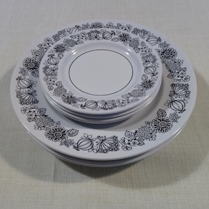 May include: A stack of white plates featuring a black floral and fruit pattern along the rim. The design includes detailed illustrations of various flowers and small round fruits arranged in a continuous circular border on each plate surface.