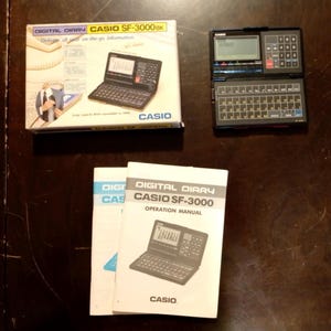 May include: A black Casio SF-3000BK digital diary with a QWERTY keyboard and LCD screen, accompanied by its original product box and an operation manual. The box features the text Digital Diary Casio SF-3000BK, and the manual is labelled Digital Diary Casio SF-3000 Operation Manual.