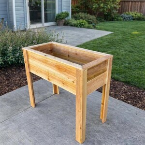 Puede incluir: Una jardinera rectangular de madera con un acabado natural en color marrón claro, que cuenta con cuatro patas verticales resistentes y un recipiente abierto profundo. La estructura está hecha de tablones horizontales con veta visible.