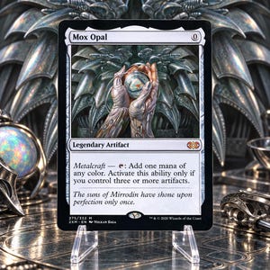 May include: A Mox Opal trading card featuring a central illustration of metallic hands holding a glowing orb. The card text includes 'Legendary Artifact', 'Metalcraft', and 'The suns of Mirrodin have shone upon perfection only once.' It is displayed on a clear stand.