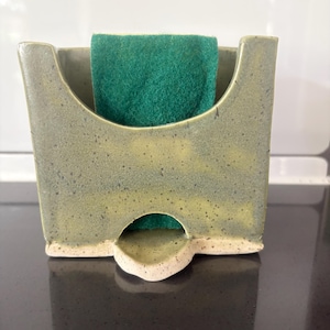 Può includere: Un porta spugna in ceramica con smalto maculato verde oliva e giallo, caratterizzato da un'ampia apertura a forma di U sulla parte superiore e un piccolo foro di drenaggio alla base. Una spugna abrasiva verde scuro è posizionata all'interno.