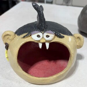 Peut inclure: Une sculpture en céramique en forme de tête ronde avec une bouche grande ouverte, présentant une peau beige, des cheveux noirs avec une mèche, des yeux blancs aux paupières violettes et deux crocs blancs. L'intérieur est peint en rouge.