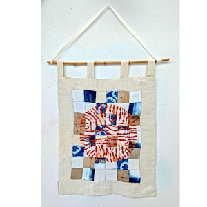 May include: A rectangular fabric wall hanging suspended from a wooden rod by four fabric tabs. The piece features a central circular motif in orange and white, surrounded by a grid of square patches in shades of blue, white, and tan.