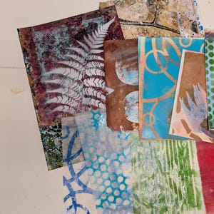 May include: A collection of rectangular paper sheets featuring various artistic prints, including a white fern silhouette on a dark textured background, blue circular patterns, green vertical stripes, and abstract gold and brown layered designs.