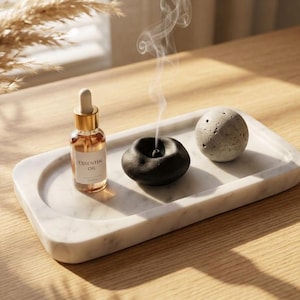 May include: A white marble tray holding a glass bottle of essential oil with a gold dropper, a black ceramic incense burner emitting a thin trail of smoke, and a textured grey concrete sphere. The items are arranged on a rectangular tray.