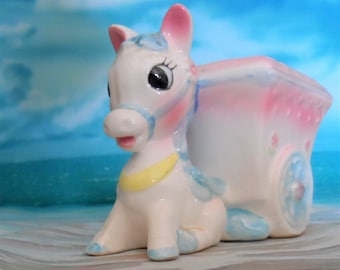 May include: A white ceramic figurine of a horse pulling a pink and blue wagon. The horse has blue eyes and a yellow collar. The wagon has a pink and blue design and a white wheel.