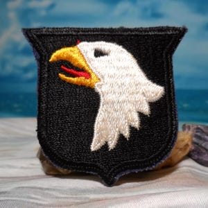 May include: Embroidered patch featuring a white eagle head with a yellow beak and red tongue, set against a black shield-shaped background. The patch has a black border and is likely an emblem or insignia.