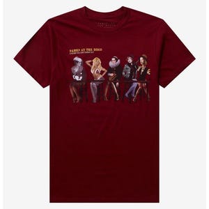 May include: A maroon short-sleeve t-shirt featuring a graphic of five figures seated at a table. Above the figures, the text reads "PANIC! AT THE DISCO A FEVER YOU CAN'T SWEAT OUT" in yellow. The figures are dressed in various costumes.