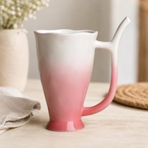 May include: A ceramic mug with a tall, tapered body and a wavy rim. The mug features a smooth ombre gradient, transitioning from white at the top to a soft pink at the base. It has a unique, elongated handle that extends upwards above the rim.