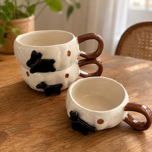 May include: Three off-white ceramic mugs with a bumpy, irregular texture. Each mug features a glossy brown handle, small brown polka dots, and a raised black animal figurine attached to the side. Two mugs are stacked vertically, and one sits beside them.
