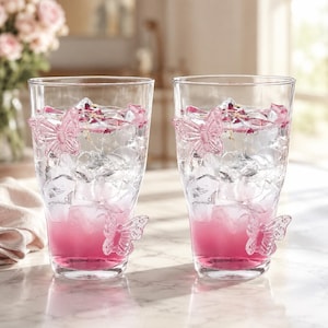 May include: Two clear glass tumblers filled with ice and a pink liquid. Each glass features two raised, three-dimensional butterfly accents on the exterior surface, one near the top rim and one near the base, crafted from clear glass.