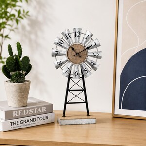 May include: A tabletop clock shaped like a windmill with a circular face featuring black numbers one through twelve. The clock has silver metal blades, a brown center, and a black metal tower base resting on a rectangular silver metal stand.
