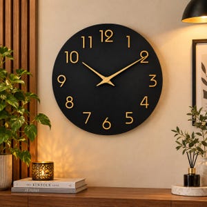 May include: A circular wall clock with a matte black face and raised gold-coloured numerals from one to twelve. The clock features two slender gold-coloured hands positioned to indicate the time, centred on the dark, solid circular background.