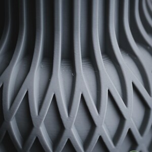 May include: A matte grey vase featuring a complex 3D printed structure with vertical ribs transitioning into a repeating diamond lattice pattern. The surface displays fine horizontal layer lines throughout the design. A small circular logo reads h3li&oslash; design.