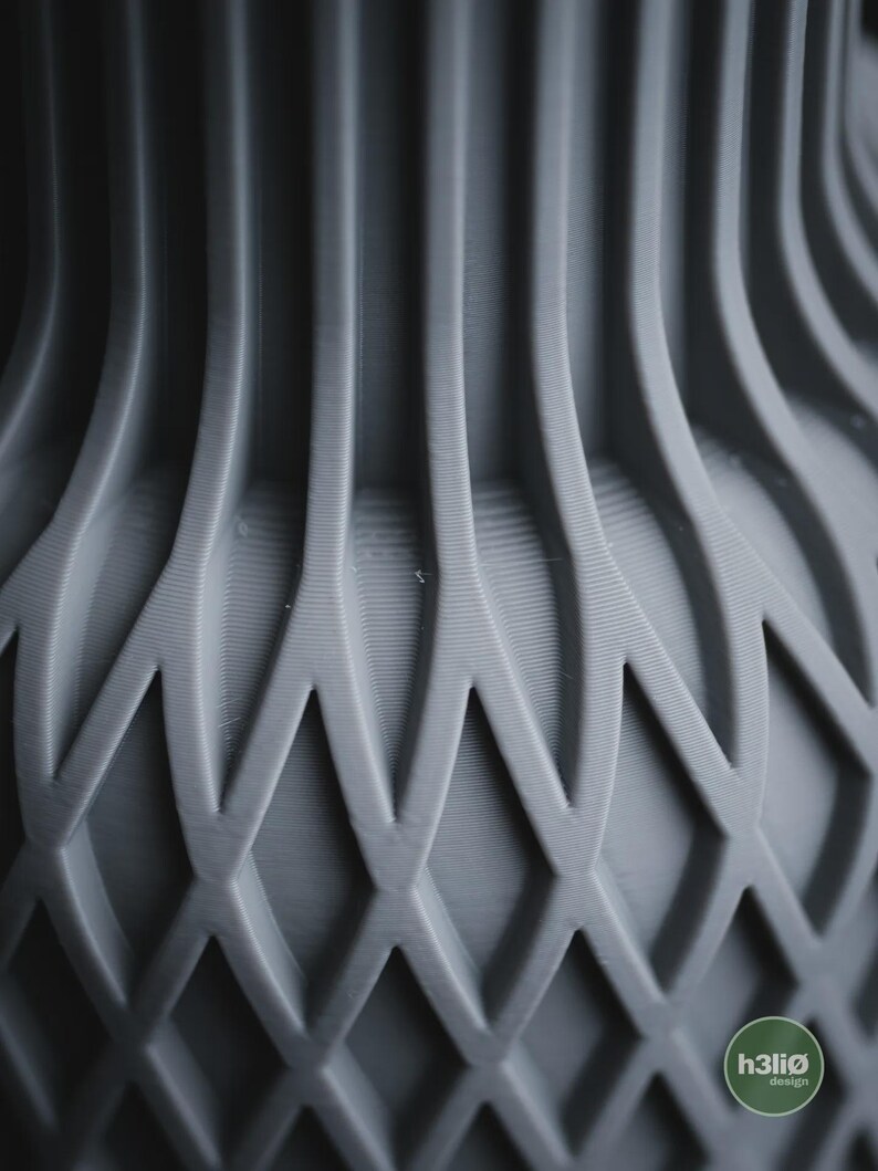 May include: A matte grey vase featuring a complex 3D printed structure with vertical ribs transitioning into a repeating diamond lattice pattern. The surface displays fine horizontal layer lines throughout the design. A small circular logo reads h3li&oslash; design.
