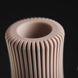 May include: A pale pink cylindrical vase featuring a series of vertical, evenly spaced ridges that extend from the top rim downwards. The top opening is circular with a smooth, thick edge. A small green circular logo with the text h3li&oslash; design is visible.