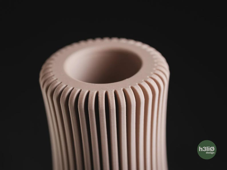 May include: A pale pink cylindrical vase featuring a series of vertical, evenly spaced ridges that extend from the top rim downwards. The top opening is circular with a smooth, thick edge. A small green circular logo with the text h3li&oslash; design is visible.