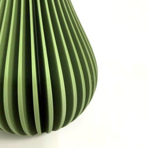 May include: A sage green vase featuring a series of vertical, accordion-style pleats that create a rhythmic, fan-like pattern around the exterior. The object has a rounded, bulbous shape with a smooth, matte finish and consistent colour throughout.
