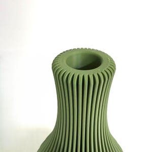 May include: A sage green vase featuring a tall, slender neck and a wide, circular opening. The exterior surface is defined by a series of vertical, parallel ridges that extend from the top rim down the length of the visible body of the object.