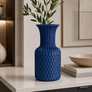 May include: A deep blue vase with a textured exterior featuring a diamond-shaped lattice pattern on the lower body and vertical fluting on the neck. The vase has a flared rim and contains a green leafy branch with several small, pointed leaves.