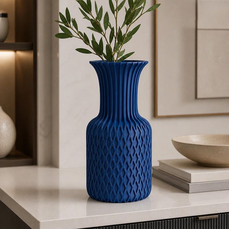 May include: A deep blue vase with a textured exterior featuring a diamond-shaped lattice pattern on the lower body and vertical fluting on the neck. The vase has a flared rim and contains a green leafy branch with several small, pointed leaves.