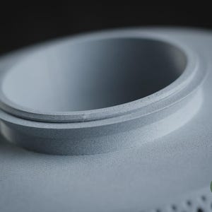 Puede incluir: Un objeto circular de color gris claro impreso en 3D con un borde central elevado y un acabado mate suave. El objeto presenta una superficie texturizada distintiva y un peque&ntilde;o logotipo circular verde en la esquina con el texto h3li&oslash; design.