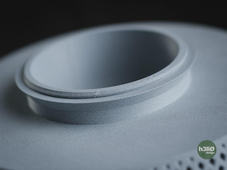 Puede incluir: Un objeto circular de color gris claro impreso en 3D con un borde central elevado y un acabado mate suave. El objeto presenta una superficie texturizada distintiva y un peque&ntilde;o logotipo circular verde en la esquina con el texto h3li&oslash; design.