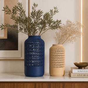 K&ouml;nnte beinhalten: Eine hohe, zylindrische dunkelblaue Vase und eine k&uuml;rzere, zylindrische beige Vase, beide mit einem vertikalen Gittermuster im oberen Bereich und einem massiven Boden. Die blaue Vase enth&auml;lt gr&uuml;ne Blattzweige, die beige Vase Trockenblumen.