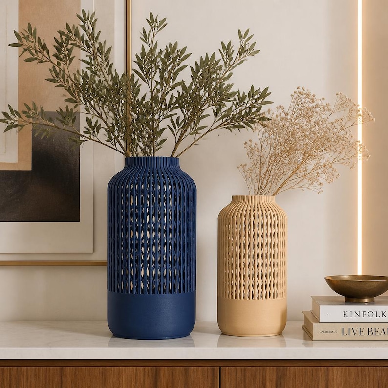 K&ouml;nnte beinhalten: Eine hohe, zylindrische dunkelblaue Vase und eine k&uuml;rzere, zylindrische beige Vase, beide mit einem vertikalen Gittermuster im oberen Bereich und einem massiven Boden. Die blaue Vase enth&auml;lt gr&uuml;ne Blattzweige, die beige Vase Trockenblumen.