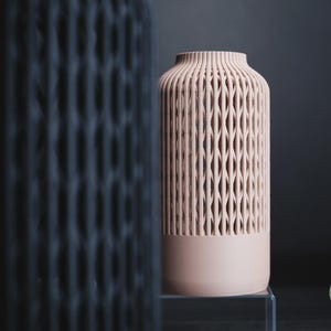 K&ouml;nnte beinhalten: Eine hellrosa zylindrische Vase mit einem vertikalen Gittermuster im oberen Bereich und einem massiven Sockel. Eine unscharfe dunkle Vase befindet sich im Vordergrund. Der Text h3li&oslash; design ist in einem gr&uuml;nen Kreis unten rechts zu sehen.