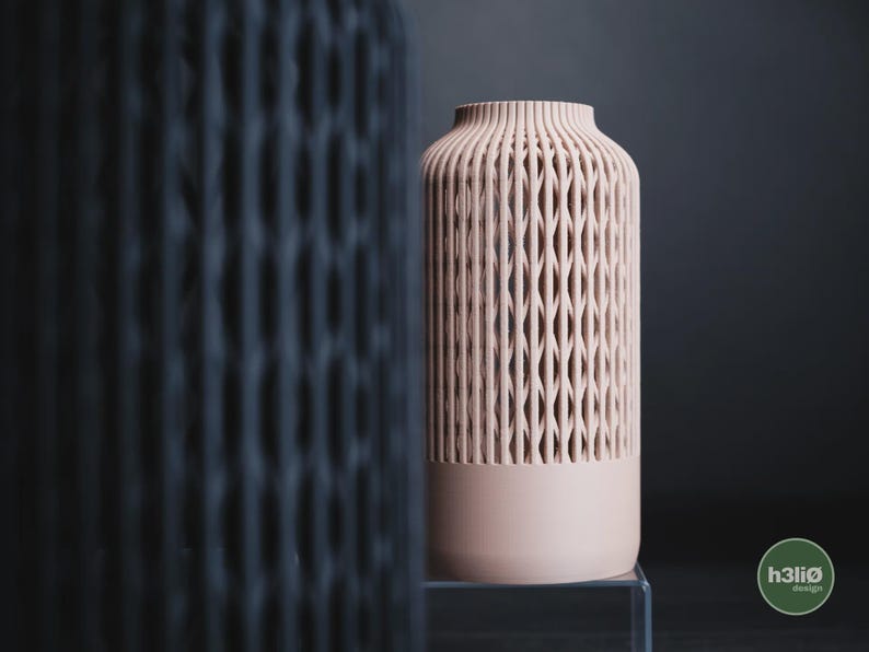 K&ouml;nnte beinhalten: Eine hellrosa zylindrische Vase mit einem vertikalen Gittermuster im oberen Bereich und einem massiven Sockel. Eine unscharfe dunkle Vase befindet sich im Vordergrund. Der Text h3li&oslash; design ist in einem gr&uuml;nen Kreis unten rechts zu sehen.