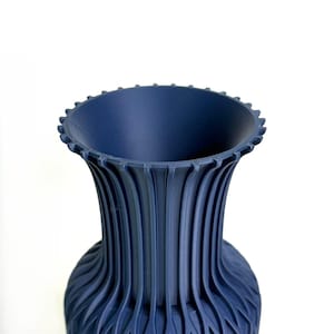 May include: A dark blue vase with a wide, flared rim featuring a series of small, evenly spaced vertical notches along the edge. The body of the vase is characterised by a repeating pattern of deep, vertical fluted ridges that extend downwards.