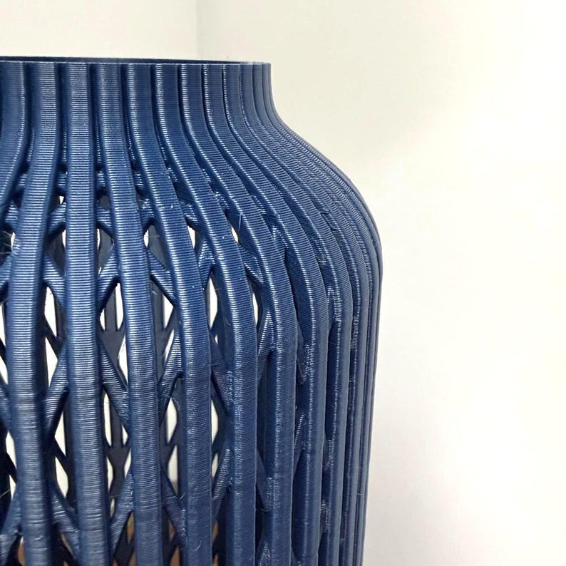 K&ouml;nnte beinhalten: Eine dunkelblaue Vase mit einer vertikal gerippten Struktur und einem komplizierten, durchbrochenen Gittermuster &uuml;ber den gesamten K&ouml;rper. Das zylindrische Objekt hat einen glatten, abgerundeten Rand und eine gleichm&auml;&szlig;ige, texturierte Oberfl&auml;che.