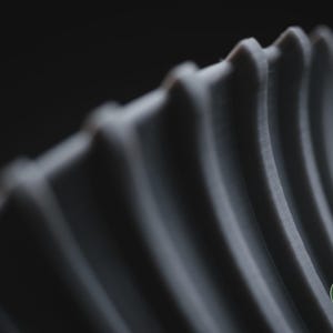 May include: A grey, three-dimensional object featuring a series of parallel, vertical, curved ridges with a textured surface. The object displays a small, circular green logo in the bottom right corner with the text h3li&oslash; design printed in white.