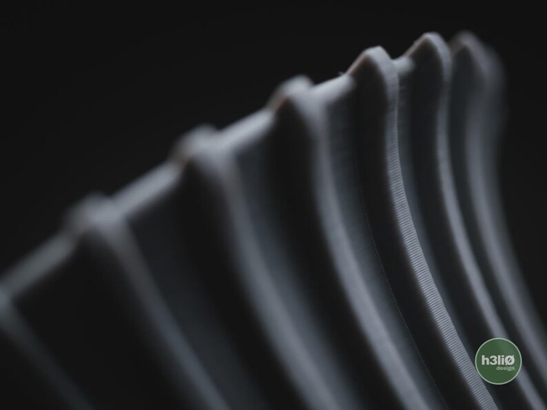 May include: A grey, three-dimensional object featuring a series of parallel, vertical, curved ridges with a textured surface. The object displays a small, circular green logo in the bottom right corner with the text h3li&oslash; design printed in white.